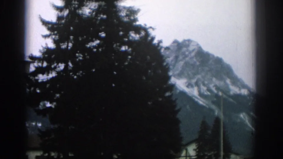 1963 Switzerland home-movie frame with a dark evergreen tree in front of a snow-marked mountain peak and pale sky.