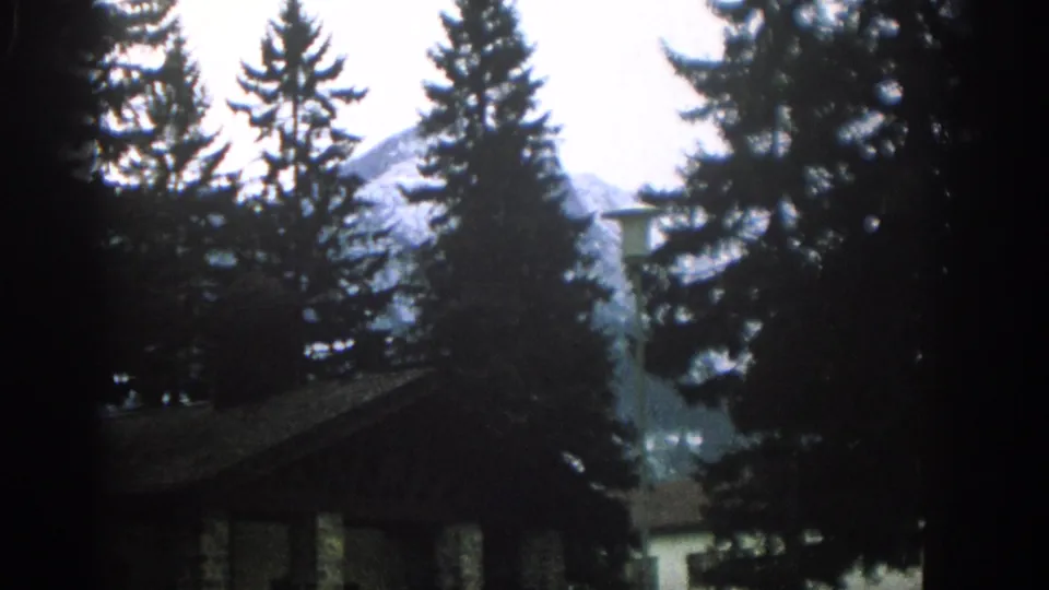 1963 Switzerland frame showing a low roofline, evergreen trees, and distant snow-marked mountains through soft 8mm color.