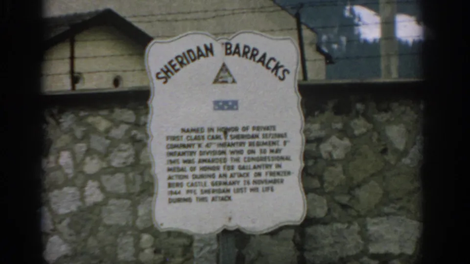 1963 Zurich 8mm frame showing the Sheridan Barracks memorial sign against a stone wall, with barbed wire and wooden siding above.