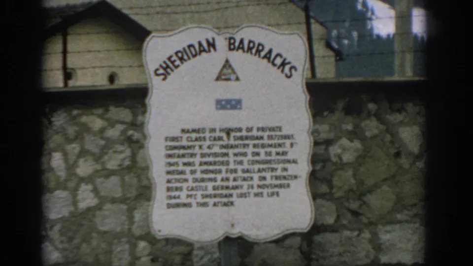 1963 Zurich home-movie frame centered on the Sheridan Barracks sign, with the memorial text visible but softened by film focus.