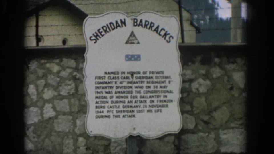 Visitor-shot 1963 frame of a Sheridan Barracks memorial plaque, with soft focus, stone masonry, and partial building detail behind it.