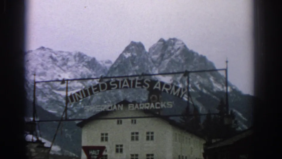 Wide 1963 Switzerland view of the United States Army Sheridan Barracks entrance sign, barracks building, and snow-capped mountains.
