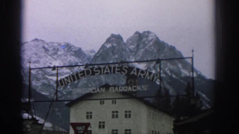 1963 8mm frame of the United States Army Sheridan Barracks entrance sign with a white building and snowy alpine peaks behind it.