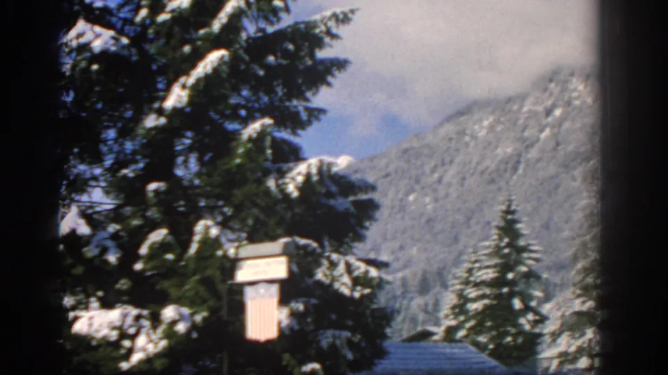 1963 8mm alpine landscape near the barracks, showing snowy evergreen trees, a small sign, and mountains partly covered by cloud.