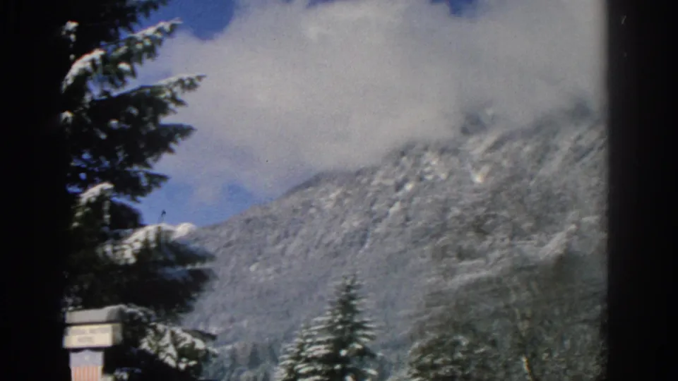 1963 8mm frame of snow-dusted alpine mountains and evergreen trees, with low clouds partly covering the peaks.