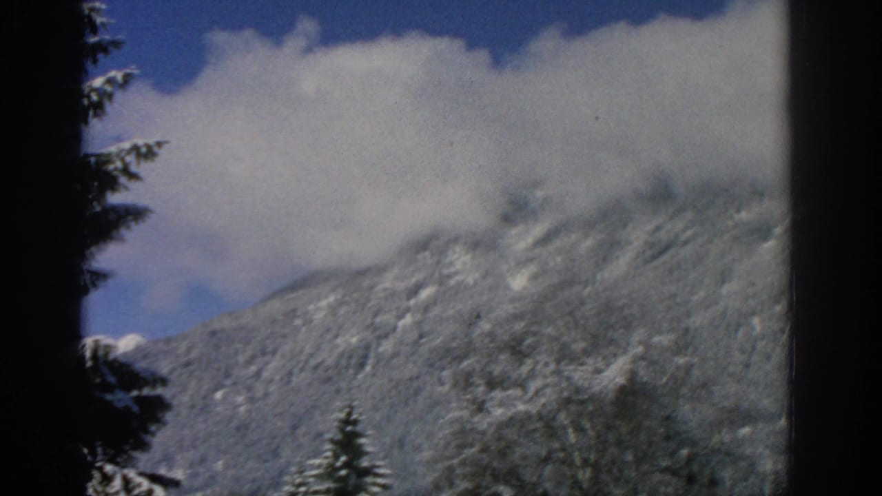 Snow-covered mountain slope beneath white clouds and blue sky, with dark evergreen edges.