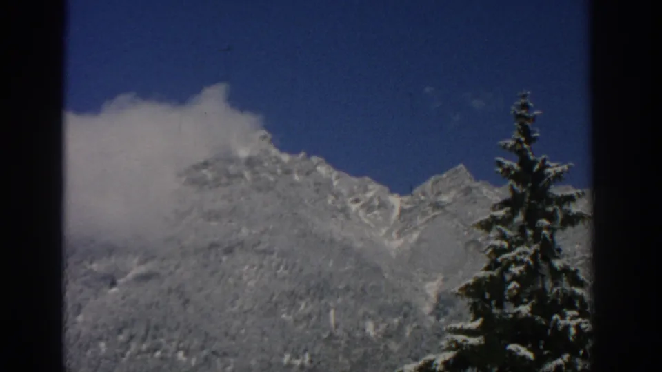 1963 8mm view near Zurich of snowy alpine peaks and clouds, with a dark evergreen tree framing the right side.