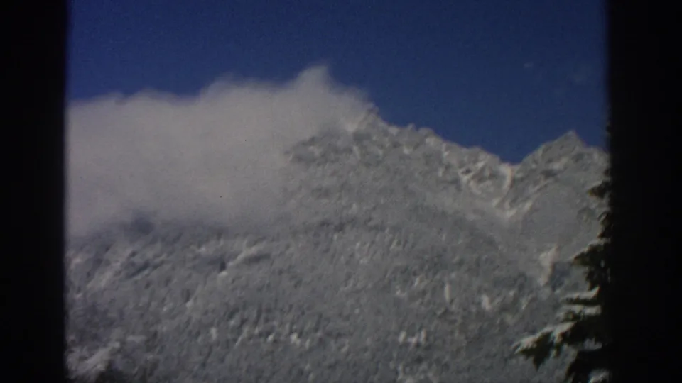 1963 Swiss 8mm frame showing a snowy mountain ridge partly veiled by a white cloud beneath a deep blue sky.