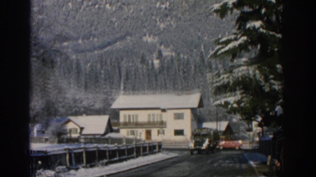 Snowy village road with houses, trees, parked vehicles, and mountains behind.