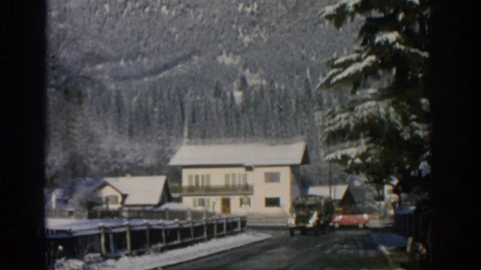 1963 Swiss 8mm view of a snow-dusted alpine village road with houses, evergreens, parked vehicles, and mountains in the background.