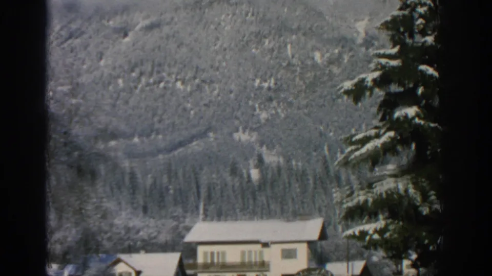 1963 Swiss 8mm frame with snow-covered mountain terrain behind village buildings and a tall evergreen at the roadside.