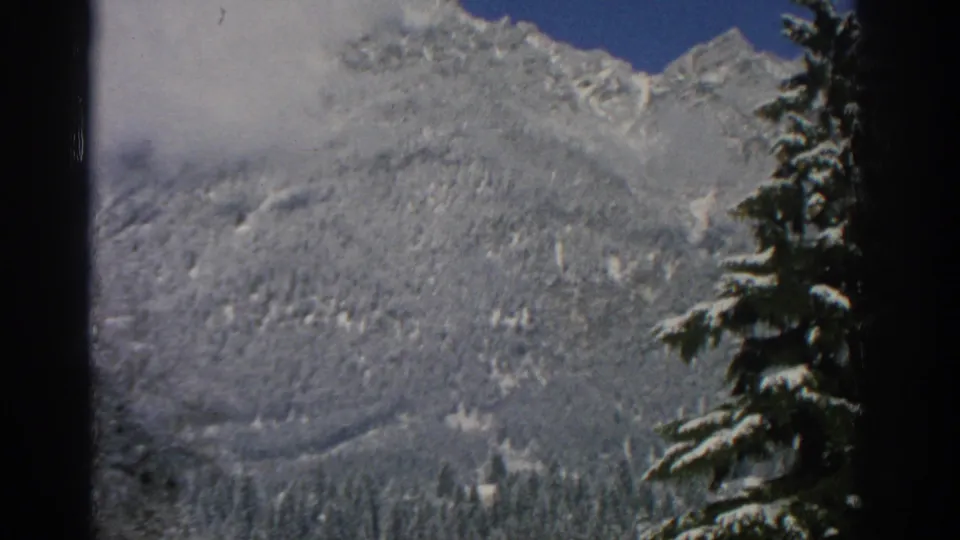 1963 Swiss 8mm view of a snow-covered mountain ridge, low cloud, blue sky, and dark evergreen branches along the frame edge.