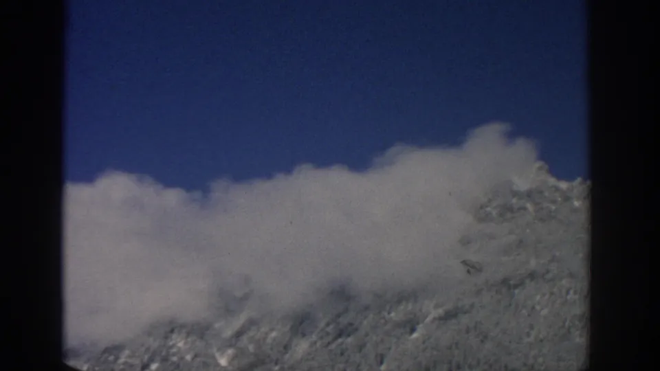 1963 Swiss alpine frame with a broad cloud bank obscuring much of the snow-covered mountain ridge beneath a clear blue sky.