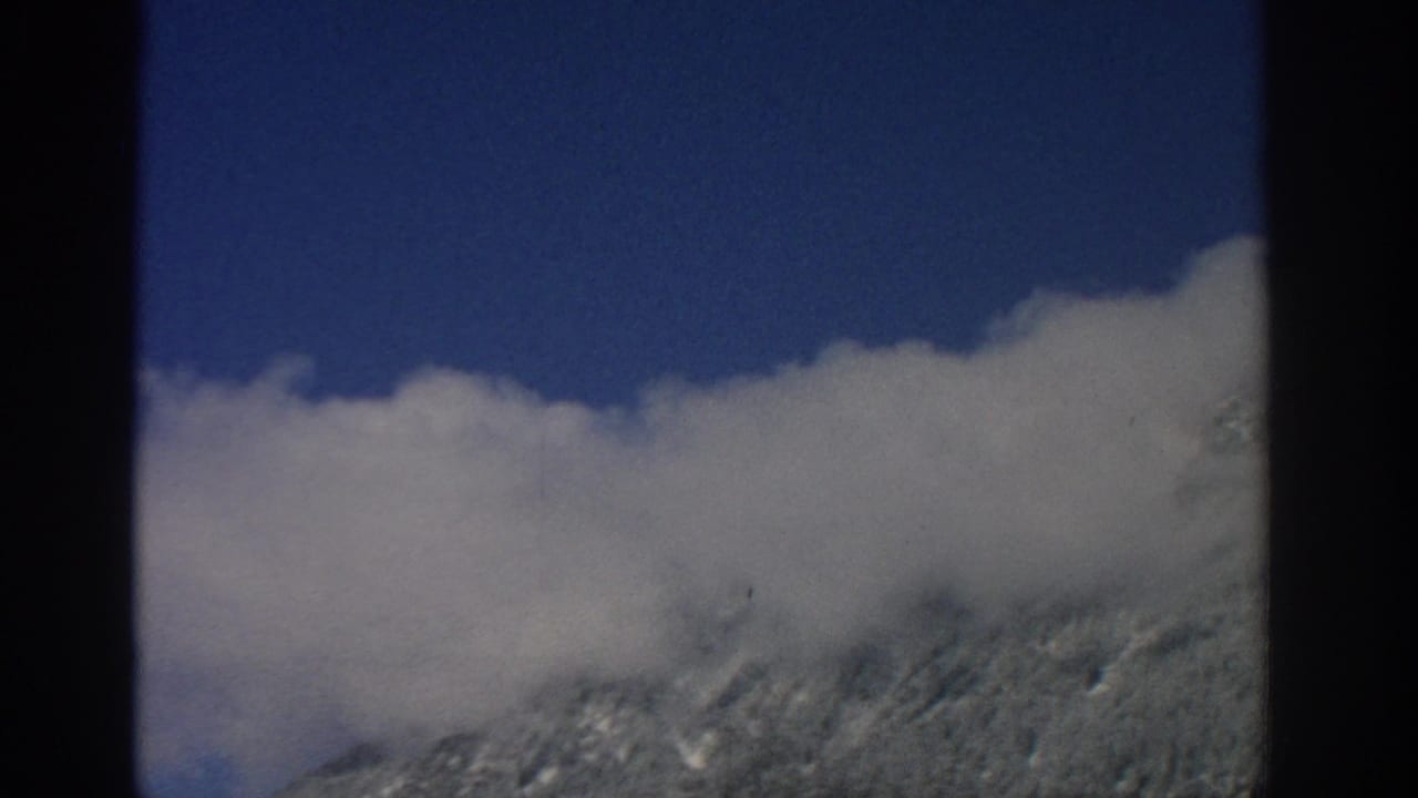 Snowy mountain slope under low white cloud and blue sky, bordered by dark film edges.