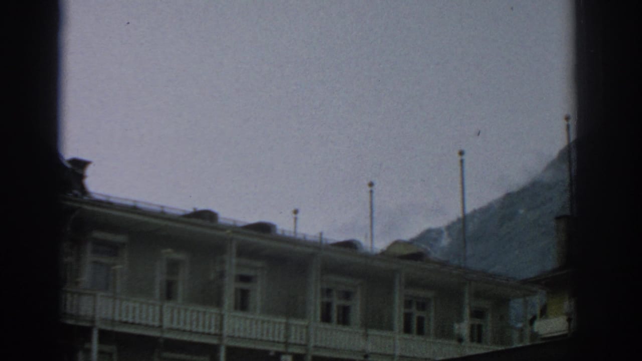 Pale lodge facade with balcony railings below a cloudy mountain backdrop.