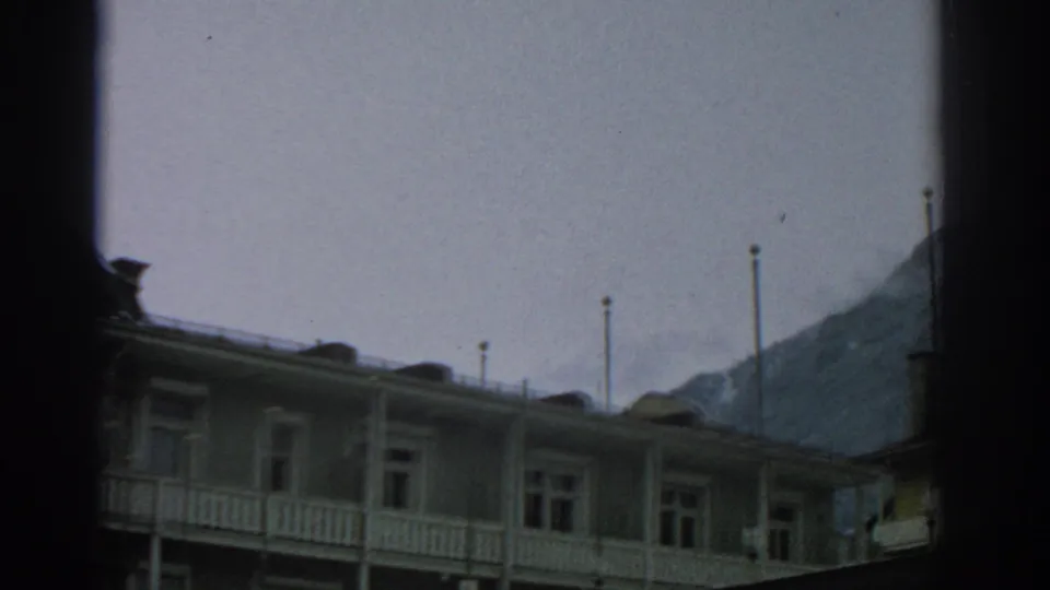 1963 8mm frame of a Swiss alpine lodge exterior with white balcony railings and a dark mountain slope behind it.