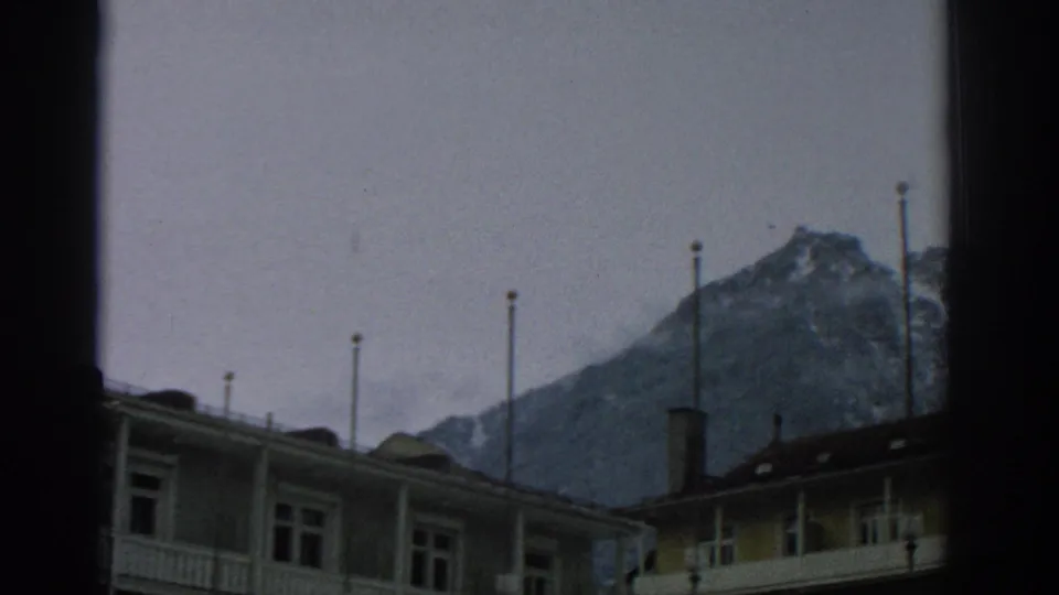 1963 Zurich-area alpine view with lodge rooflines, tall flagpoles, and snow-dusted peaks under an overcast sky.