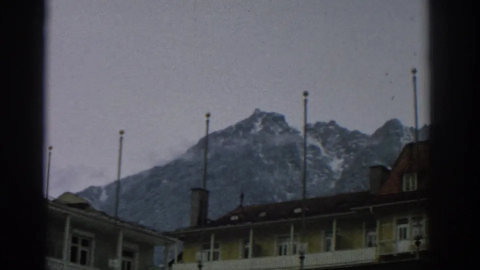 1963 8mm frame showing Swiss alpine peaks above lodge rooftops, chimneys, flagpoles, and pale balcony railings.
