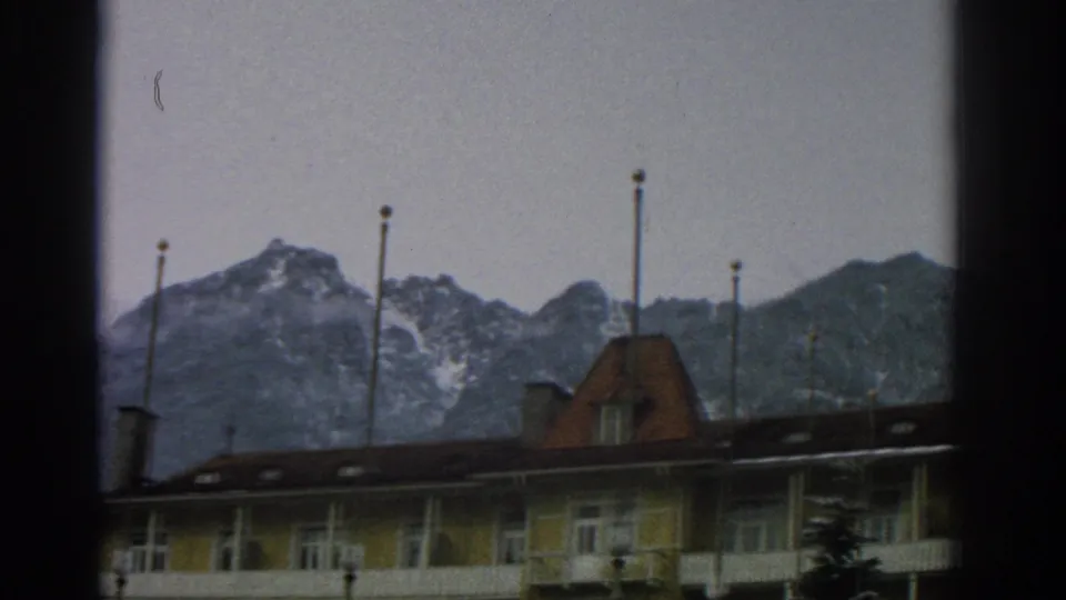 Late pan frame from 1963 Zurich-area 8mm footage, with a yellow lodge wing, balcony rail, chimneys, and alpine peaks.