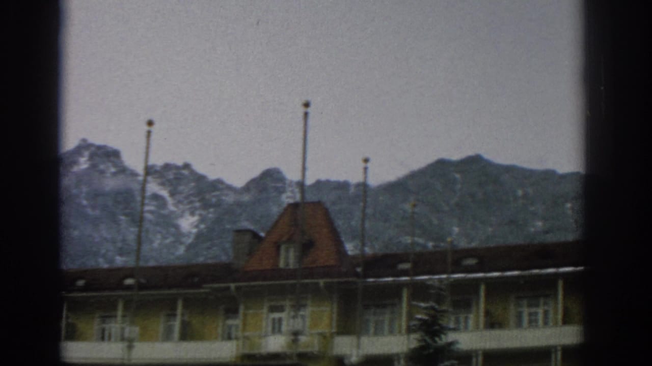 Yellow building with red-tiled roof below snow-dusted mountains, seen through dark window-frame edges.