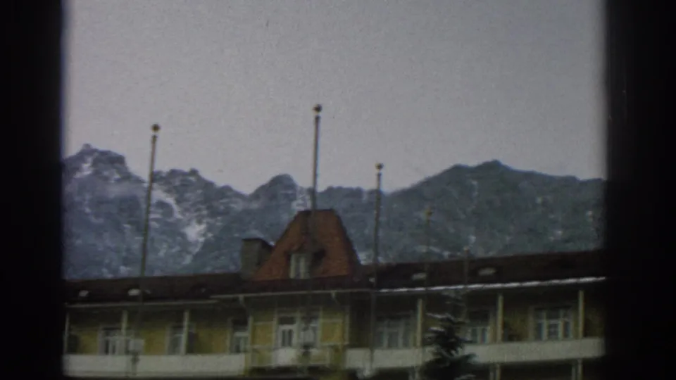 1963 Zurich home-movie frame showing a yellow alpine-style building with flagpoles and mountains behind it, partly obscured by window framing.
