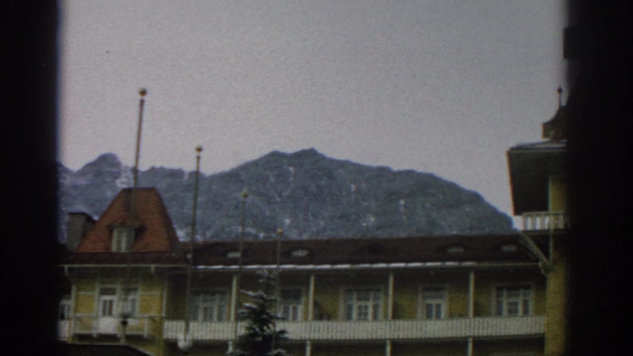 Similar mountain-backed building view with balcony railings, flagpoles, and dark vertical window framing.