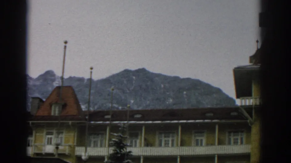 1963 Zurich exterior view of a yellow building and balcony railings beneath a dark roof, with alpine hills behind and window edges in the foreground.