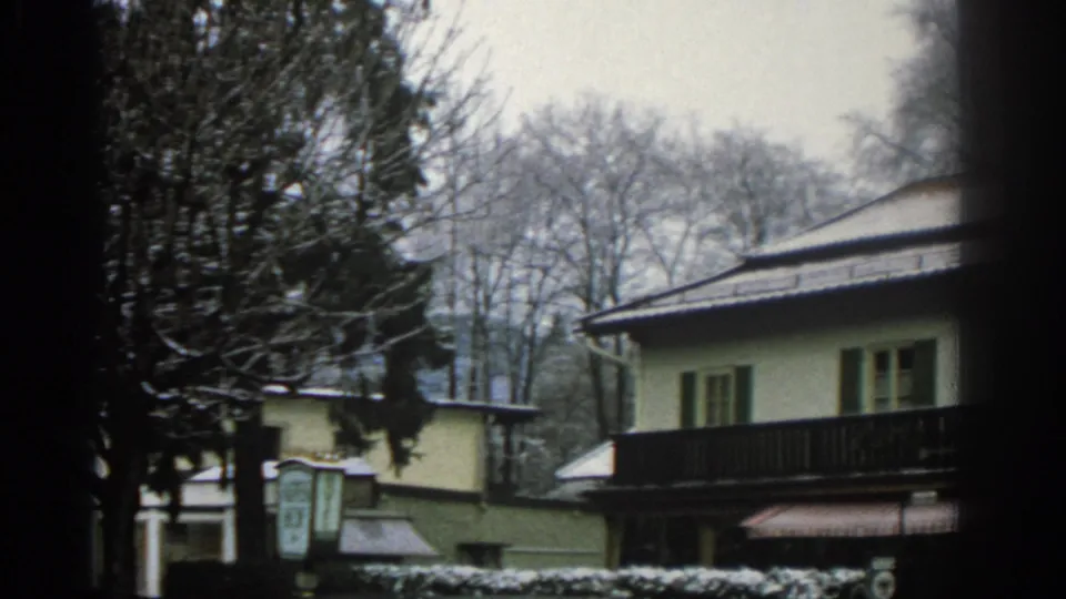 1963 Zurich home-movie view of a pale residential building with a dark balcony, snowy roof edges, bare trees, and a small sign-like panel at left.