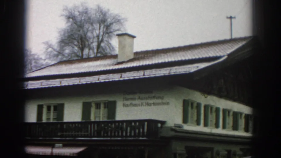 1963 Zurich 8mm still showing the building corner with snow on the roof, a wooden balcony, green shutters, and German wall text.