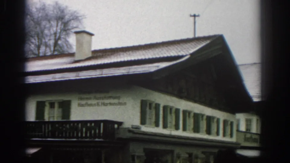 1963 Zurich frame with the snowy roofline, white facade, green shutters, balcony railing, German text, and a dark timber gable.