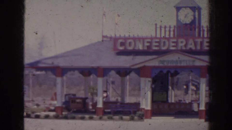 1962 Florida 8mm frame showing a closer view of the attraction pavilion, large CONFEDERATE lettering, a clock tower, and small train cars beneath.