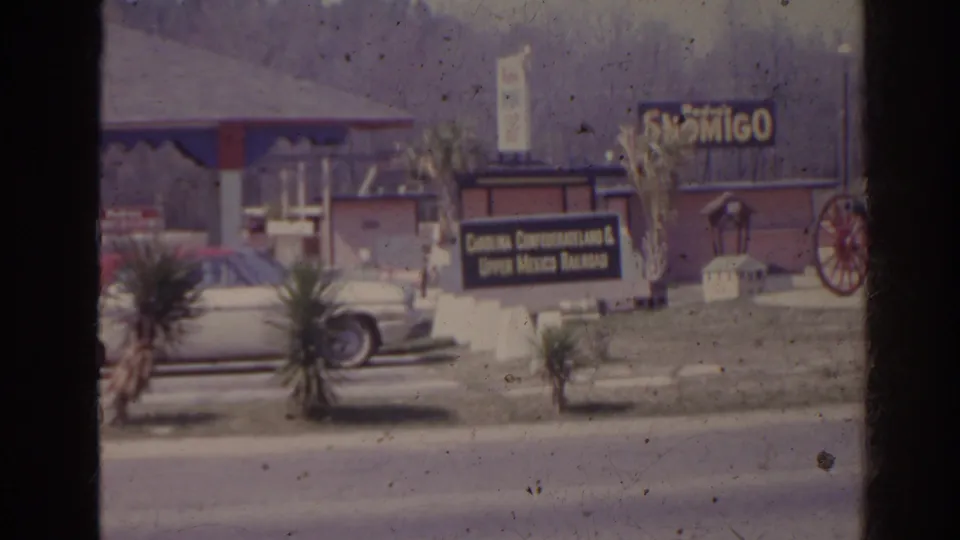 1962 Florida home-movie frame with roadside Confederateland signage, a parked car, planting, and wagon wheel detail outside the attraction.