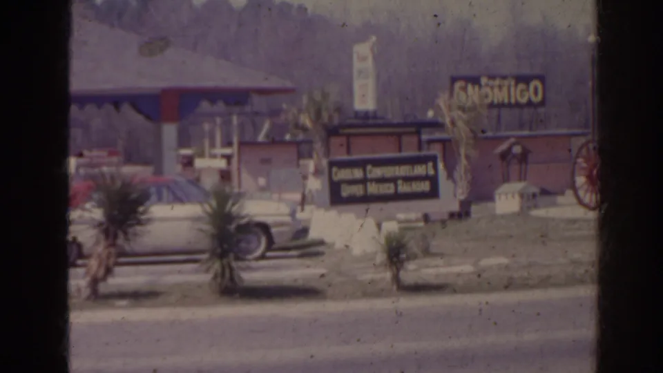 1962 Florida roadside view with a gas station canopy, parked car, Son Migo sign, and foreground sign for dining and Mexican bazaar.