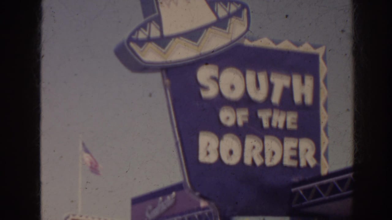 Large blue and yellow South of the Border sign in the 1962 Florida roadside scene, with a sombrero-shaped top and sky behind it.