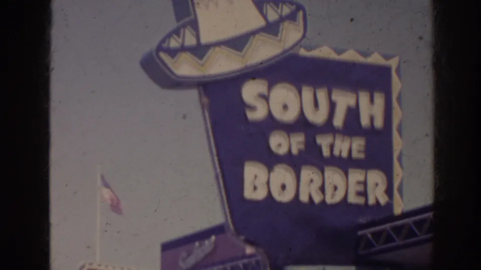 Large blue and yellow South of the Border sign in the 1962 Florida roadside scene, with a sombrero-shaped top and sky behind it.