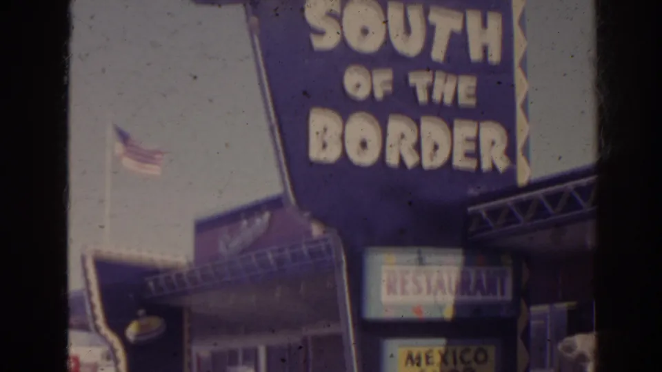1962 Florida roadside storefront with South of the Border sign above restaurant and Mexico Shop signs, parked cars, and a flag nearby.