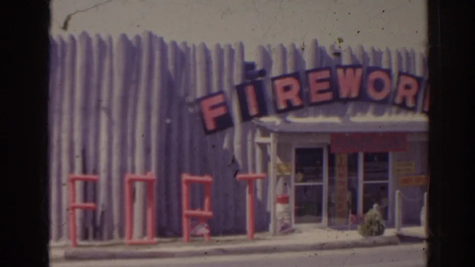 1962 Florida frame of a roadside fireworks-style shop with partial FIREWORK lettering, FORT facade letters, doorway, and small signs near the entrance.