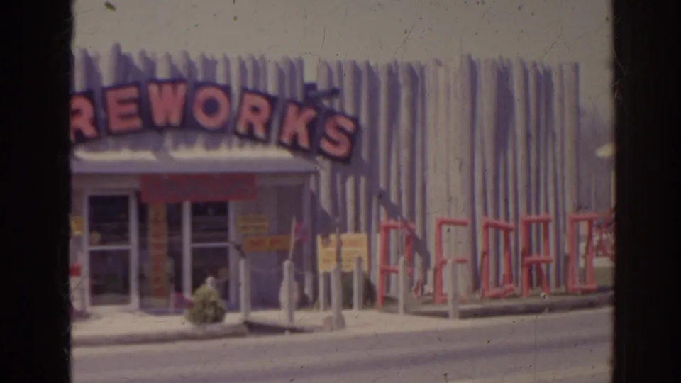 1962 Florida frame with a passing car crossing in front of the fireworks shop and its red arched sign.