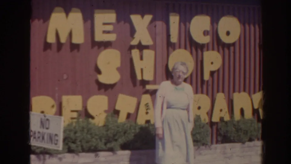 1962 Florida home-movie frame with a woman outside Mexico Shop Restaurant, framed by shrubs, signage, and a no-parking sign.
