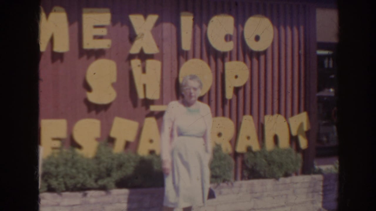 Elderly woman standing before a red wall with large Mexico Shop Restaurant lettering.