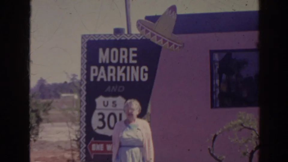 An elderly woman stands by a pink roadside building beside a More Parking sign with a U.S. route shield and sombrero topper.