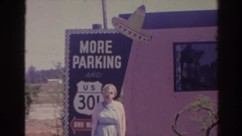 The camera frames the blue More Parking sign, U.S. route shield, sombrero decoration, pink wall, and partial view of the woman.