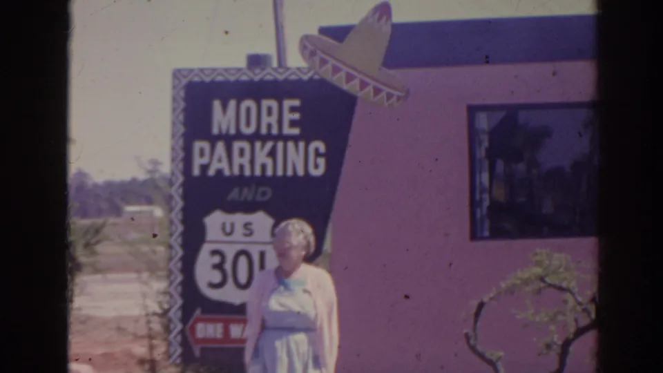 An elderly woman in a light dress stands below the More Parking sign, with the pink exterior wall, window, and small tree visible.