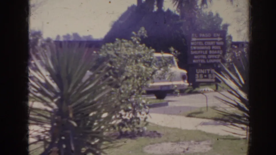 Florida 1962 home-movie frame with the El Paso Inn sign, a white sedan, motel landscaping, and spiky plants framing the parking area.