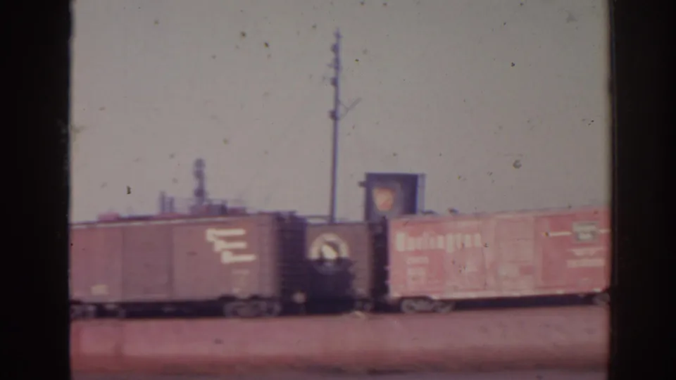 1962 Florida home-movie frame showing freight cars on rail tracks, with industrial buildings, poles, and hazy sky in the background.