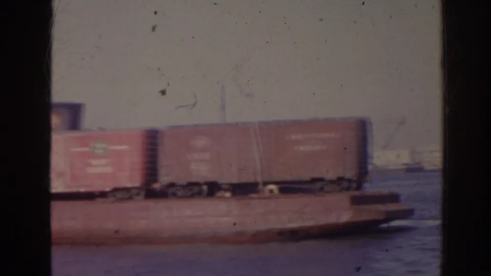 1962 Florida 8mm frame showing brown and red boxcars on a low barge with open water visible beyond them.