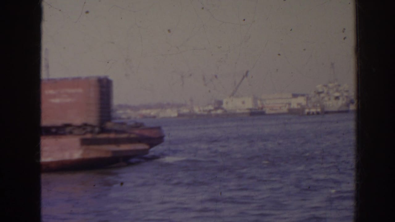 1962 Florida harbor view with water, a barge or docked platform at left, distant industrial waterfront, and dark window-like edges framing the shot.