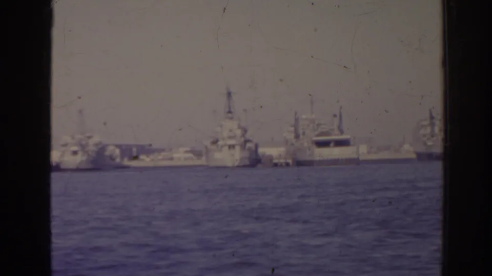 1962 Florida 8mm frame showing distant naval ships lined along a harbor, viewed across blue water with soft focus and film grain.