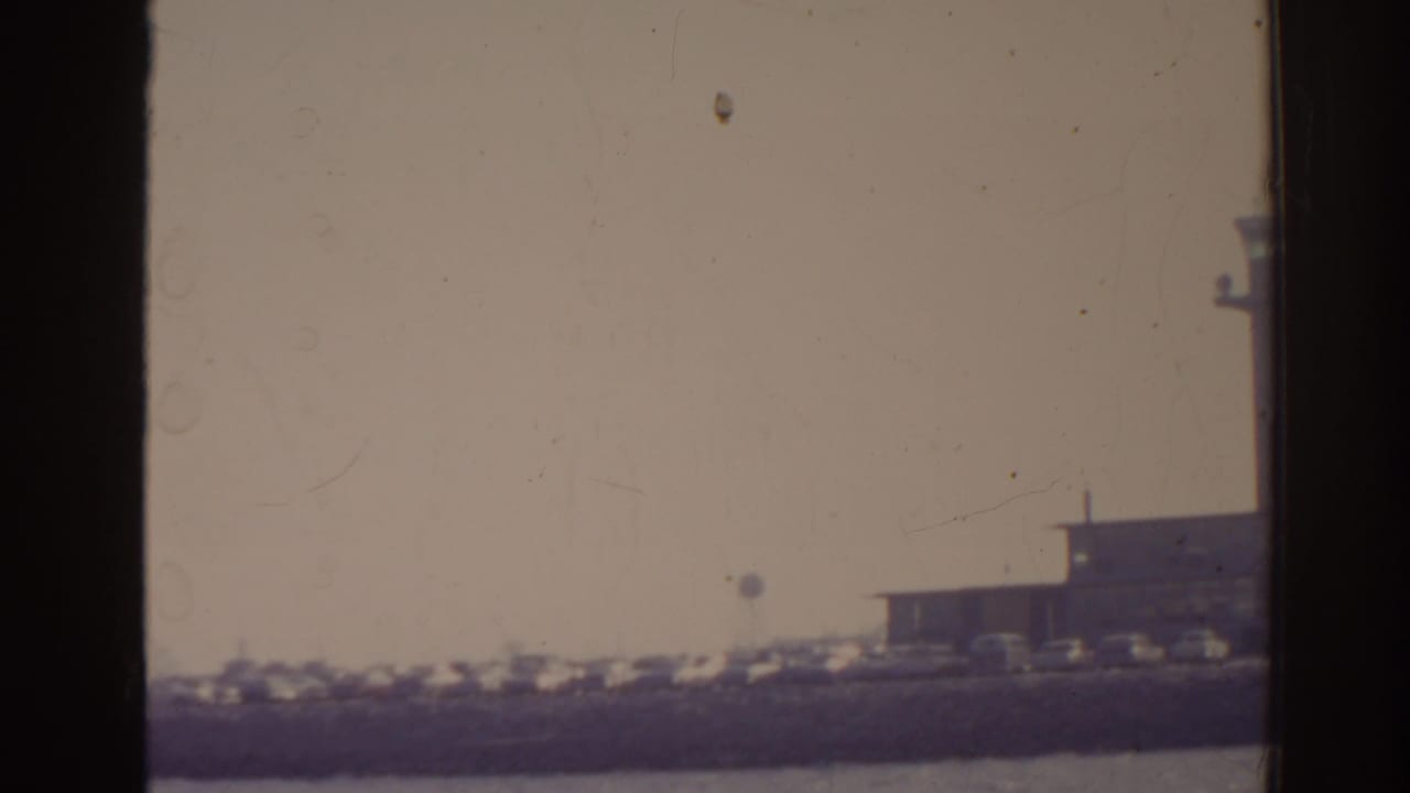 1962 Florida 8mm frame showing a hazy distant airport terminal, shoreline parking, and water in the foreground.