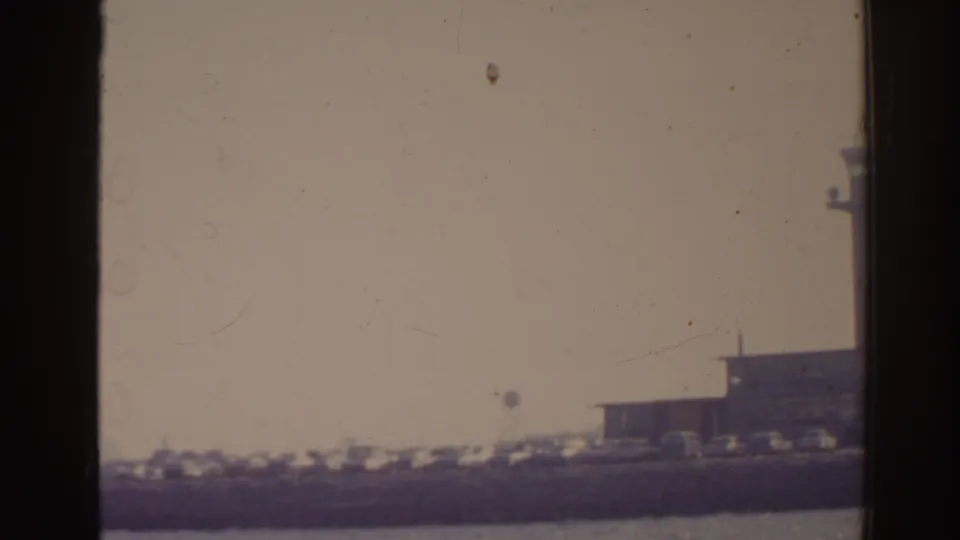 1962 Florida 8mm frame showing a hazy distant airport terminal, shoreline parking, and water in the foreground.